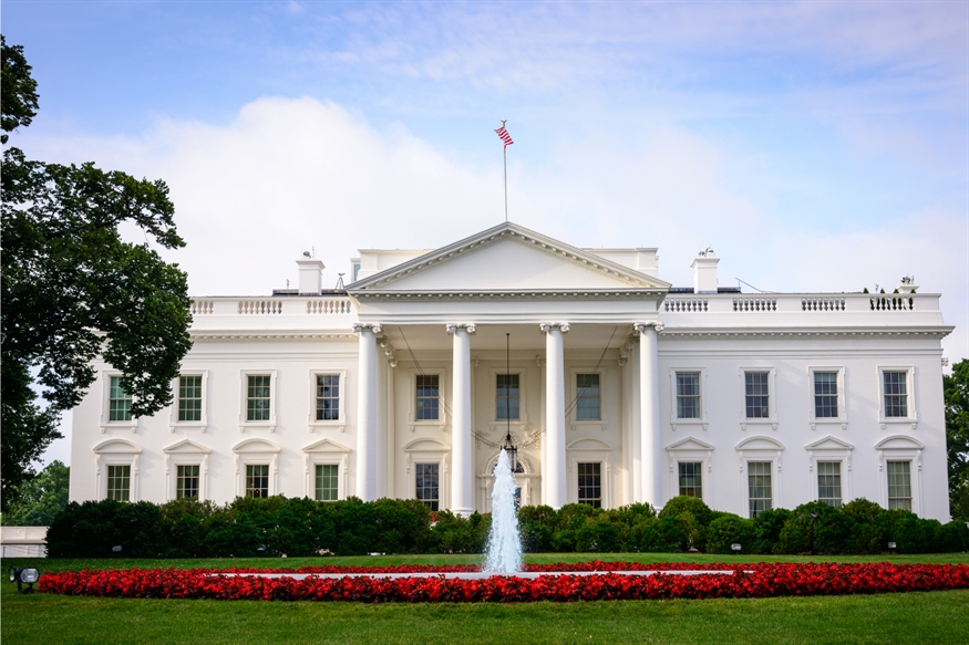 [ai] The White House, a large white-painted neoclassical building, with a fountain and well-maintained garden featuring red flowers in the foreground. The American flag is displayed on the roof.